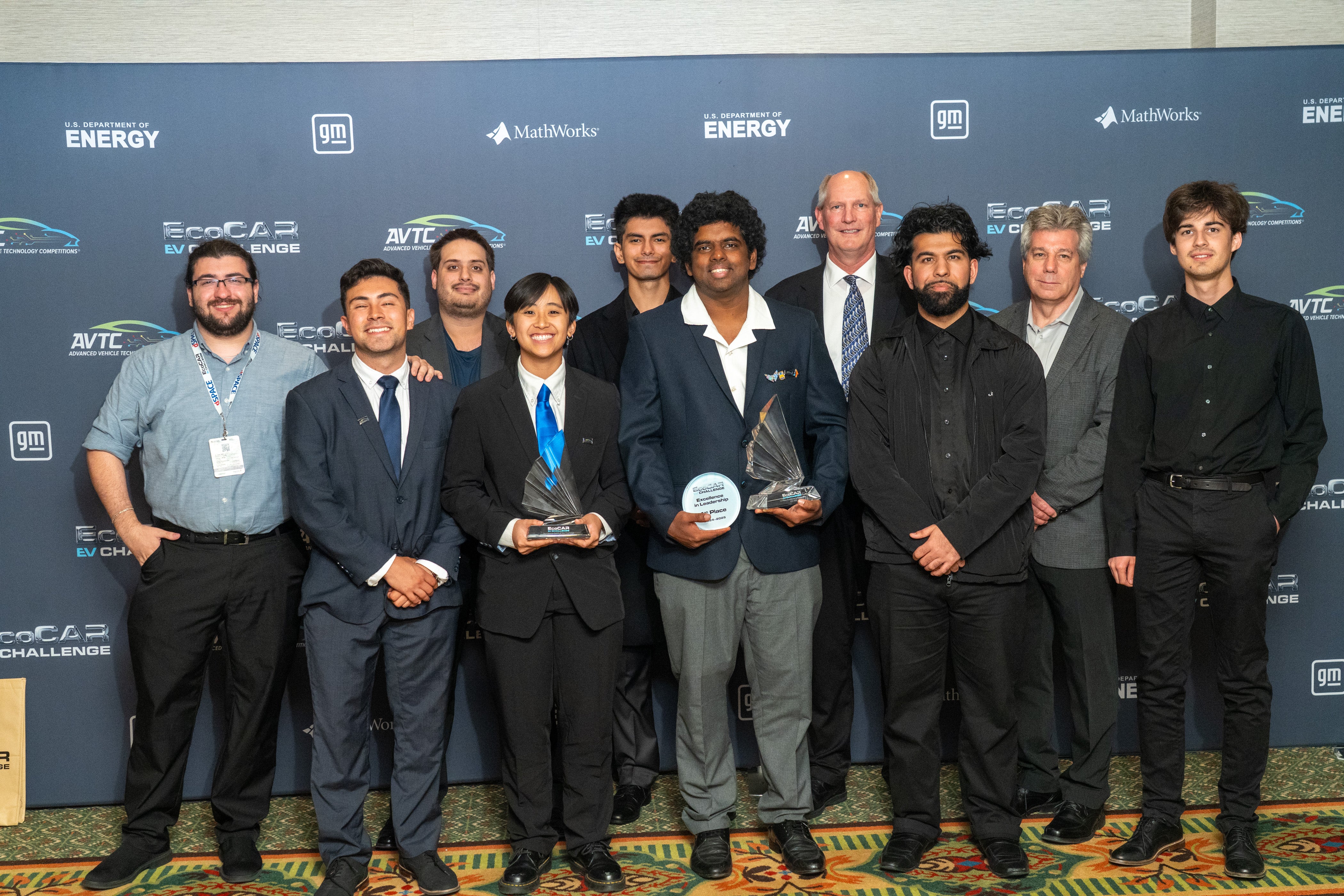 UC Riverside EcoCAR EV Challenge team members and mentors pose together on stage, holding multiple trophies in front of a competition backdrop featuring logos from the U.S. Department of Energy, General Motors, and MathWorks.