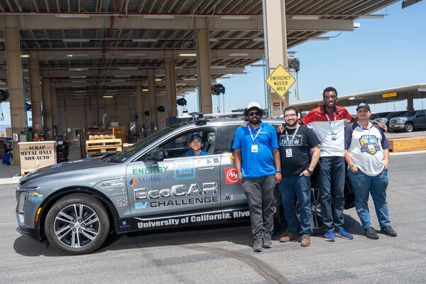 Five UC Riverside EcoCAR EV Challenge team members pose next to a gray Cadillac LYRIQ covered with sponsor logos, including the U.S. Department of Energy, General Motors, and MathWorks. One team member sits in the driver’s seat smiling, while the others stand beside the vehicle in an outdoor automotive facility.
