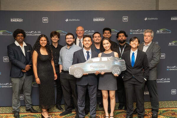 EcoCAR EV Challenge team group photo in front of event backdrop, with members smiling and holding a car-shaped sign.