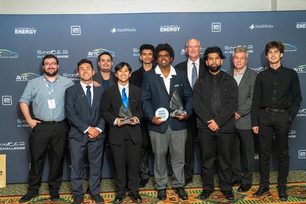 A group of UC Riverside EcoCAR EV Challenge team members and mentors pose together on the award stage, holding trophies for their achievements. They stand in front of a branded backdrop featuring logos from the U.S. Department of Energy, General Motors, and MathWorks.